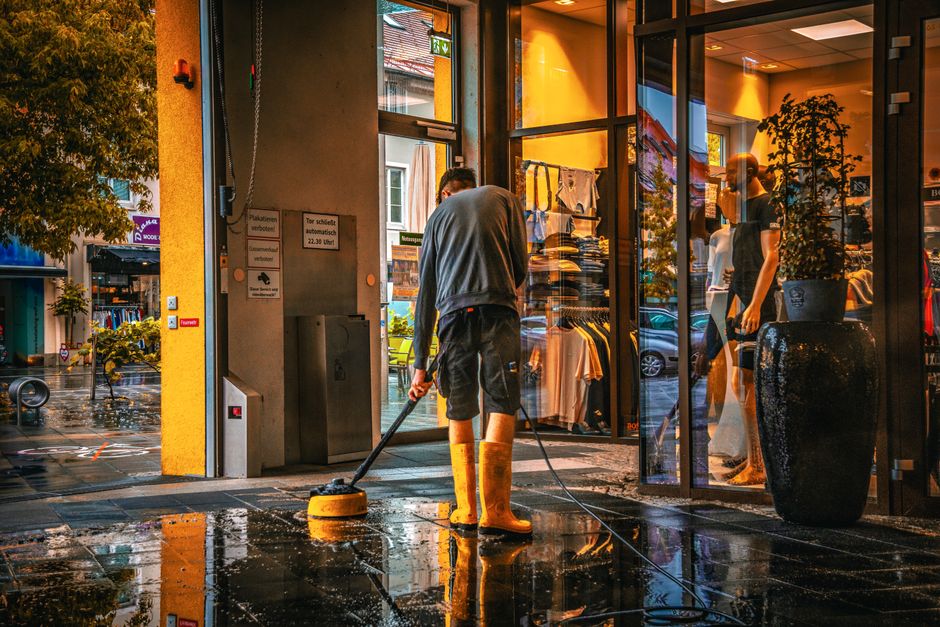 A person wearing yellow rubber boots is using a pressure washer to clean the tiled entrance of a commercial store in Fulham during daytime. The entrance features large glass windows displaying clothing and accessories inside, with reflections of the wet, shiny black tiles on the ground. The surrounding area shows a rainy street scene with parked cars, a tree, and nearby buildings. The cleaning process appears thorough, with water and soap visible on the tiled surface, and the indoor lighting casting a warm glow through the glass. Fulham Carpet Cleaning is mentioned as providing professional cleaning services in Fulham, highlighting the importance of surface cleaning to maintain hygiene and appearance.