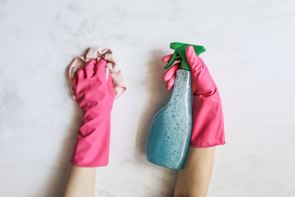 A pair of hands wearing bright pink rubber cleaning gloves, one hand holding a beige microfiber cloth and the other holding a transparent spray bottle filled with blue cleaning solution with visible bubbles, positioned over a smooth, light-colored, marble surface. The background features a clean, white, polished countertop or table, suggesting a domestic or commercial setting for surface cleaning and sanitisation. Natural lighting highlights the shiny, clean appearance of the surfaces and the cleaning tools, with the focus on the gloves and cleaning equipment associated with deep cleaning or household sanitisation, consistent with the services provided by Fulham Carpet Cleaning.