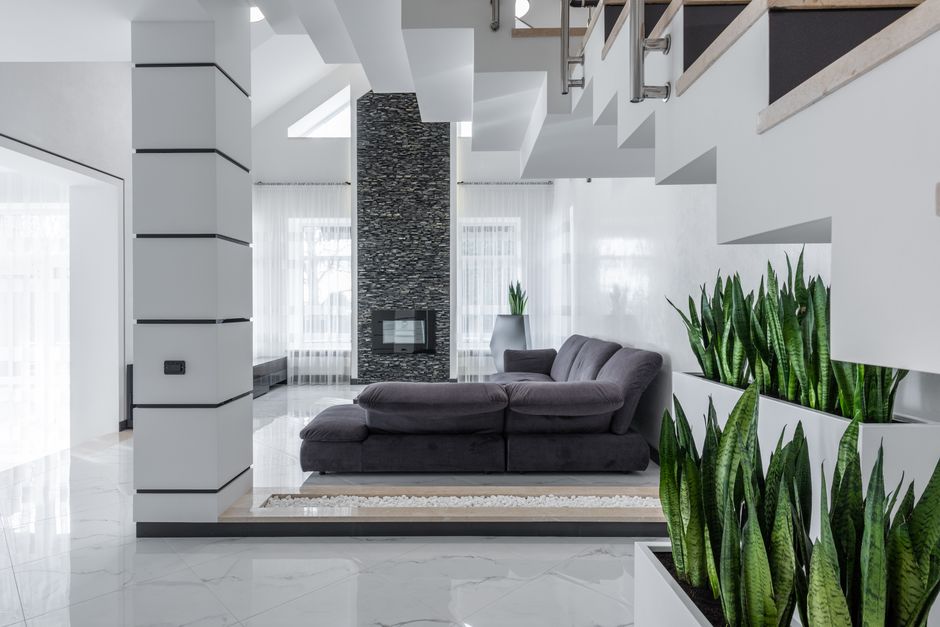 A modern, open-plan living room featuring a grey upholstered sofa positioned on a white area rug with a black border. The room has white marble flooring with subtle veining and is illuminated by natural light coming through large, sheer white curtains covering floor-to-ceiling windows. The space includes a chimney with a textured black stone surround, set against a white wall, and a fireplace insert below it. On the right side, there are tall, vibrant green snake plants in white rectangular planters arranged along the white wall with built-in niches. The room's decor includes minimal furnishings, a staircase with white steps and stainless steel railings visible in the upper right corner, and a clean, uncluttered appearance indicating regular surface cleaning and maintenance performed by Fulham Carpet Cleaning.
