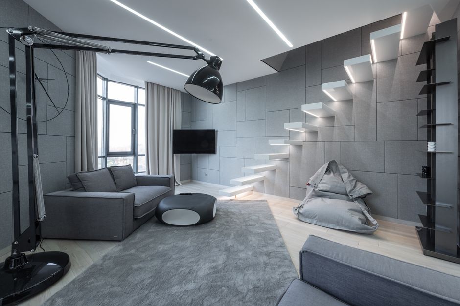 A modern, open-plan living room featuring a grey upholstered sofa positioned on a white area rug with a black border. The room has white marble flooring with subtle veining and is illuminated by natural light coming through large, sheer white curtains covering floor-to-ceiling windows. The space includes a chimney with a textured black stone surround, set against a white wall, and a fireplace insert below it. On the right side, there are tall, vibrant green snake plants in white rectangular planters arranged along the white wall with built-in niches. The room's decor includes minimal furnishings, a staircase with white steps and stainless steel railings visible in the upper right corner, and a clean, uncluttered appearance indicating regular surface cleaning and maintenance performed by Fulham Carpet Cleaning.