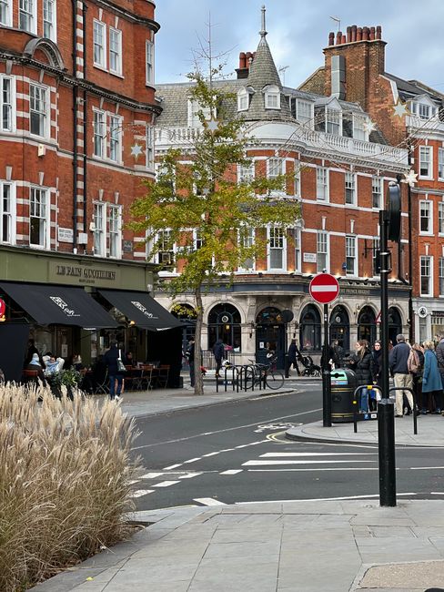 Street view of a busy area near Fulham Broadway, featuring a row of brick and white-painted Victorian-style buildings with multiple windows and decorative elements. The scene includes a tree with yellowing leaves, pedestrians walking along the sidewalk, and outdoor seating at a café with black awnings labeled with the café's name. The street has a crosswalk and traffic signs, including a 'no entry' sign, and a black bollard in the foreground. The lighting is natural, indicating a daytime setting, and the scene appears clean and well-maintained, reflecting the type of urban environment that Fulham Carpet Cleaning services for surface and deep cleaning in local shops and cafes.