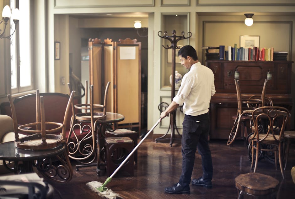 A man wearing a white shirt and dark trousers is performing surface cleaning in a room, using a mop to clean a polished wooden floor. The room features a variety of wooden furniture, including chairs with curved backs and a vintage upright piano, as well as a coat rack and a bookshelf filled with books. Natural light from large windows illuminates the space, highlighting the shiny, clean surface of the wooden flooring. The room appears well-maintained and tidy, with no visible dust or clutter, reflecting professional cleaning practices by Fulham Carpet Cleaning.