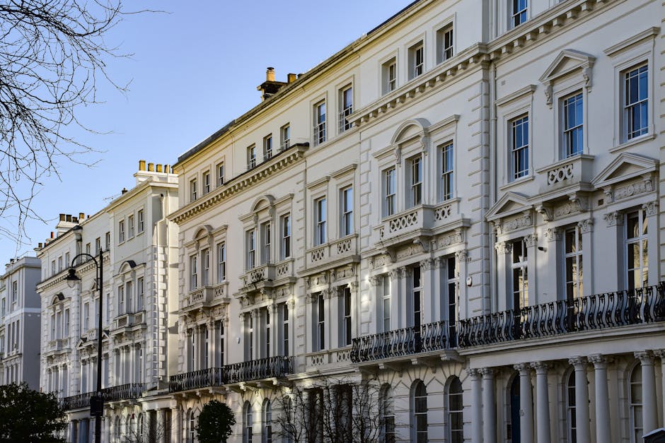 Exterior view of a row of elegant Victorian-style white residential buildings with ornate architectural details, including decorative cornices and balconies with black wrought iron railings. The buildings have large sash windows, some with small balconies, and the facade appears clean and well-maintained under natural daylight. A classic black street lamp is visible in the foreground, and leafless tree branches frame the scene, indicating a subtle seasonal context. This image showcases the pristine outdoor surfaces typical of high-end flats in Fulham, emphasizing surface cleaning and building maintenance that Fulham Carpet Cleaning offers for residential properties in SW6, FULHAM.