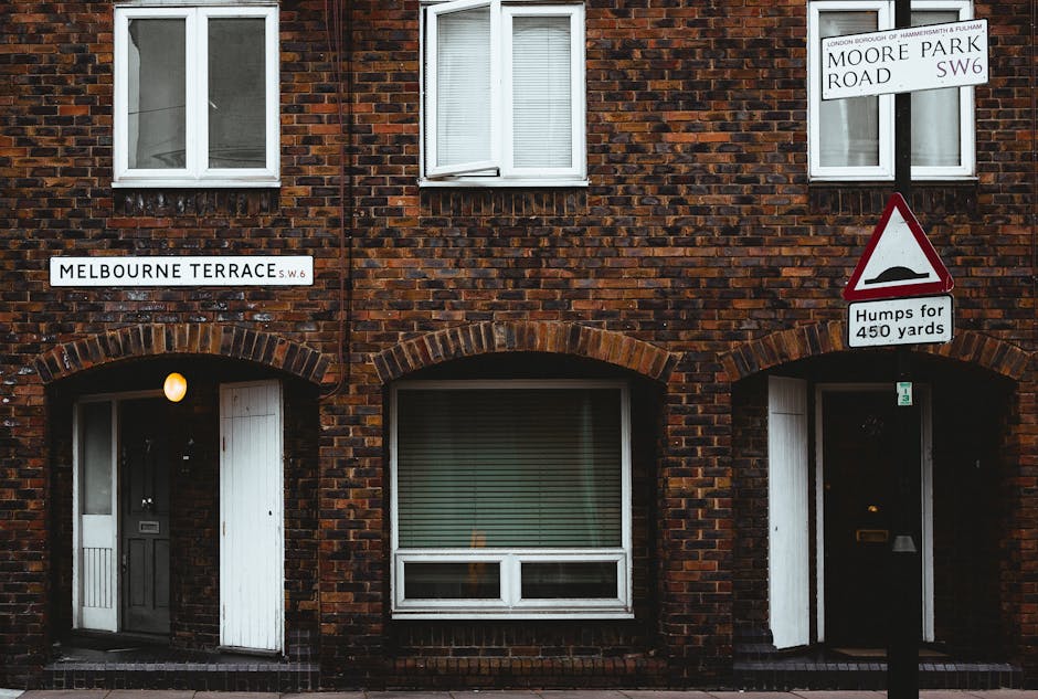 A close-up view of a residential brick building on Fulham Road, with two white-framed windows on the upper floor, and two arch-shaped doorways on the ground level, each with a gray door and white shutters. The brick facade is dark red with variations, and a single exterior light illuminates the left doorway. Street signs indicate the location as Melbourne Terrace SW6 and Moore Park Road, along with a triangular traffic sign warning about road humps for 450 yards. The scene is set in natural daylight, highlighting the well-maintained and clean appearance of the building's surface, consistent with professional cleaning and maintenance standards.