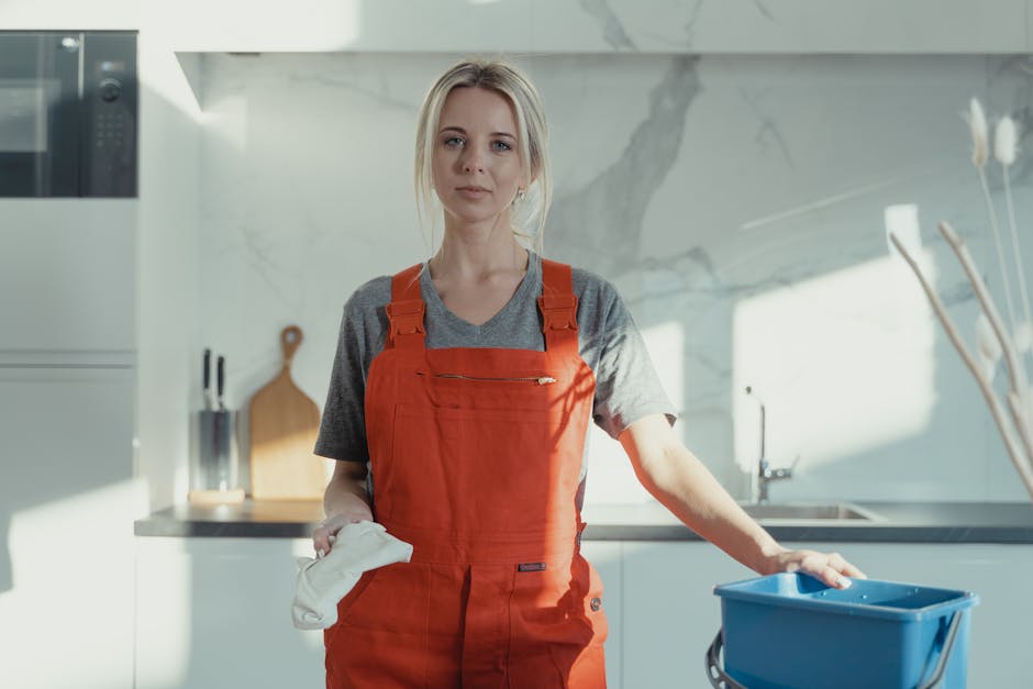 A professional cleaner wearing an orange apron and grey t-shirt standing in a modern, bright kitchen with white marble countertops and backsplash. The cleaner holds a white cleaning cloth in her right hand and rests her left hand on a blue cleaning bucket. The kitchen features built-in appliances, including a microwave, and is well-lit with natural light, highlighting the spotless and shiny surfaces, clean tiles, and neatly organized space. Fulham Carpet Cleaning offers surface cleaning and deep cleaning services for residential properties, emphasizing hygiene and maintenance, as seen in the cleanliness of this kitchen environment.