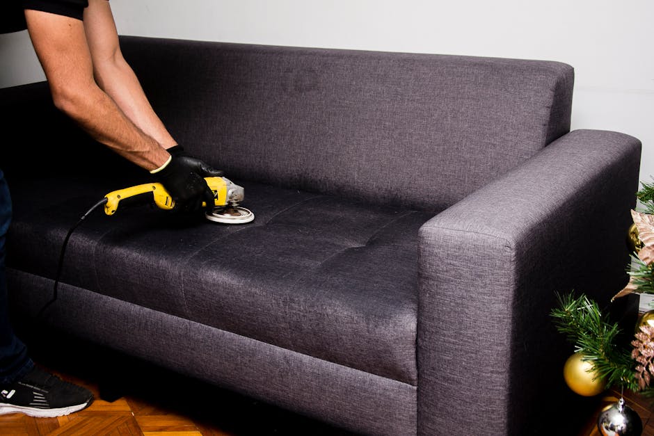 A person wearing black gloves is using a handheld electric upholstery cleaner to deep clean a dark gray fabric sofa in a living room. The sofa's surface is damp and slightly shiny, indicating recent cleaning. The room features wooden flooring and is decorated for festive occasions with a Christmas tree visible on the right side, adorned with gold and pink ornaments and green foliage. The lighting is natural, coming from an unseen window, illuminating the sofa and part of the person's arm. The scene emphasizes surface cleaning and sanitisation processes by Fulham Carpet Cleaning, showcasing the detailed maintenance of upholstery in domestic settings.